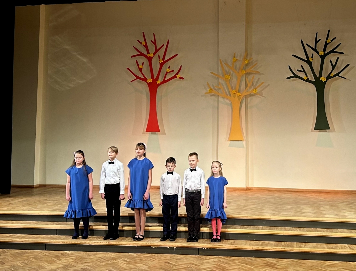 6 bērni - 3 meitenes zilās kleitās, 3 zeņi baltos kreklos un melnās biksēs dzied  uz skatuves, fonā dekorācijās krāsaini koki 02.2024.