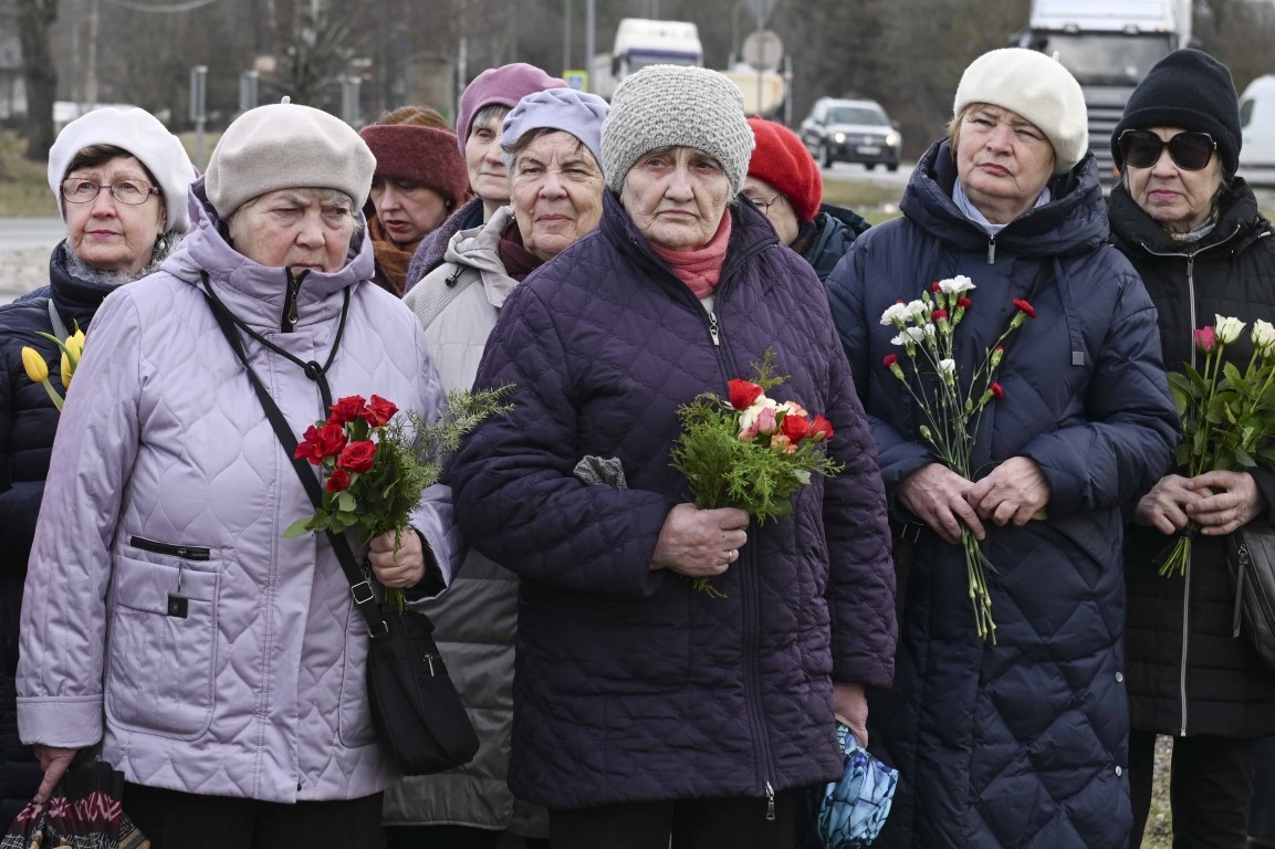 Vecāka gadagājuma cilvēki stāv ar sarkaniem ziediem