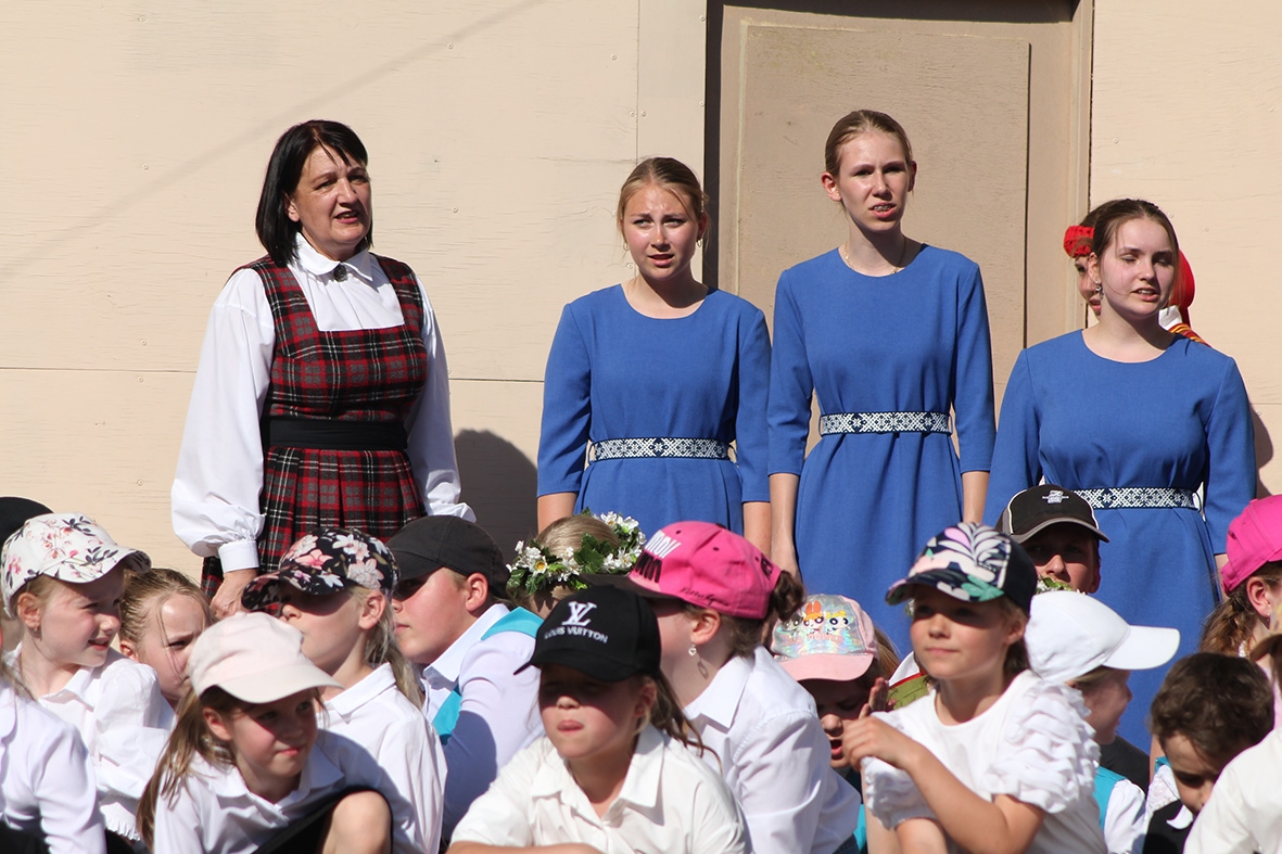 Skatītāji, 2 meitenes zilos tērpos un skolotāja stāv pasākumā Sadziedāsim Suntažos! 24.05.2024.