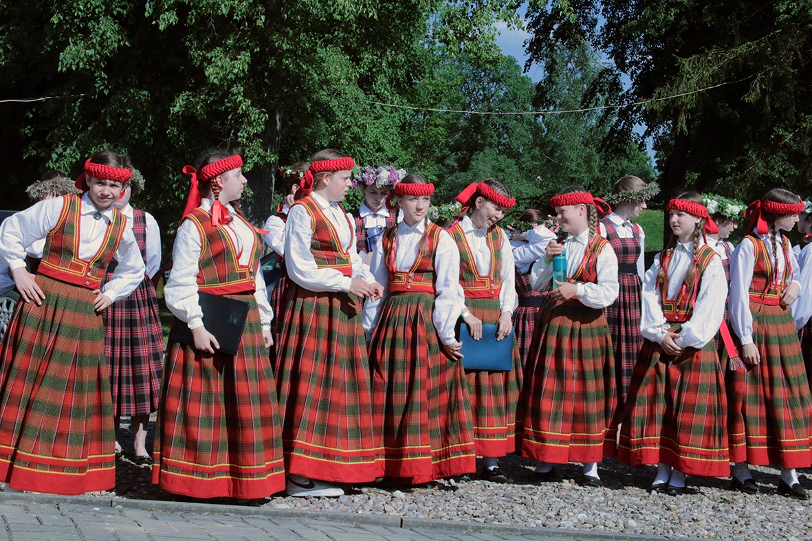 Kora dalībnieki rūtainos tautas tērpos stāv pasākumā Sadziedāsim Suntažos! 24.05.2024.