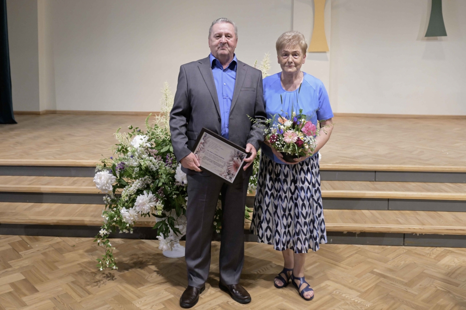 Sieviete zilganā kleitā ar ziediem, vīrietis uzvalkā ar pateicības rakstu Ogres novada Zelta kāzu pāris 31.05.2024