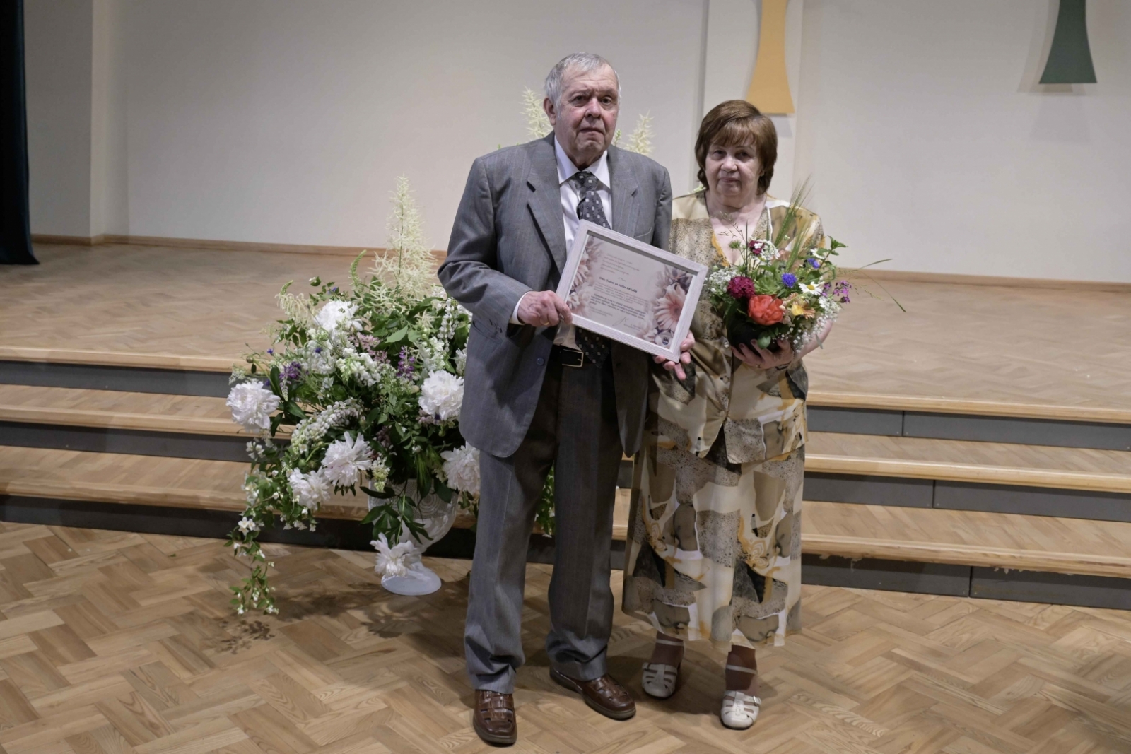 Sieviete sūnu krāsas kleitā ar ziediem, vīrietis uzvalkā ar pateicības rakstu Ogres novada Zelta kāzu pāris 31.05.2024