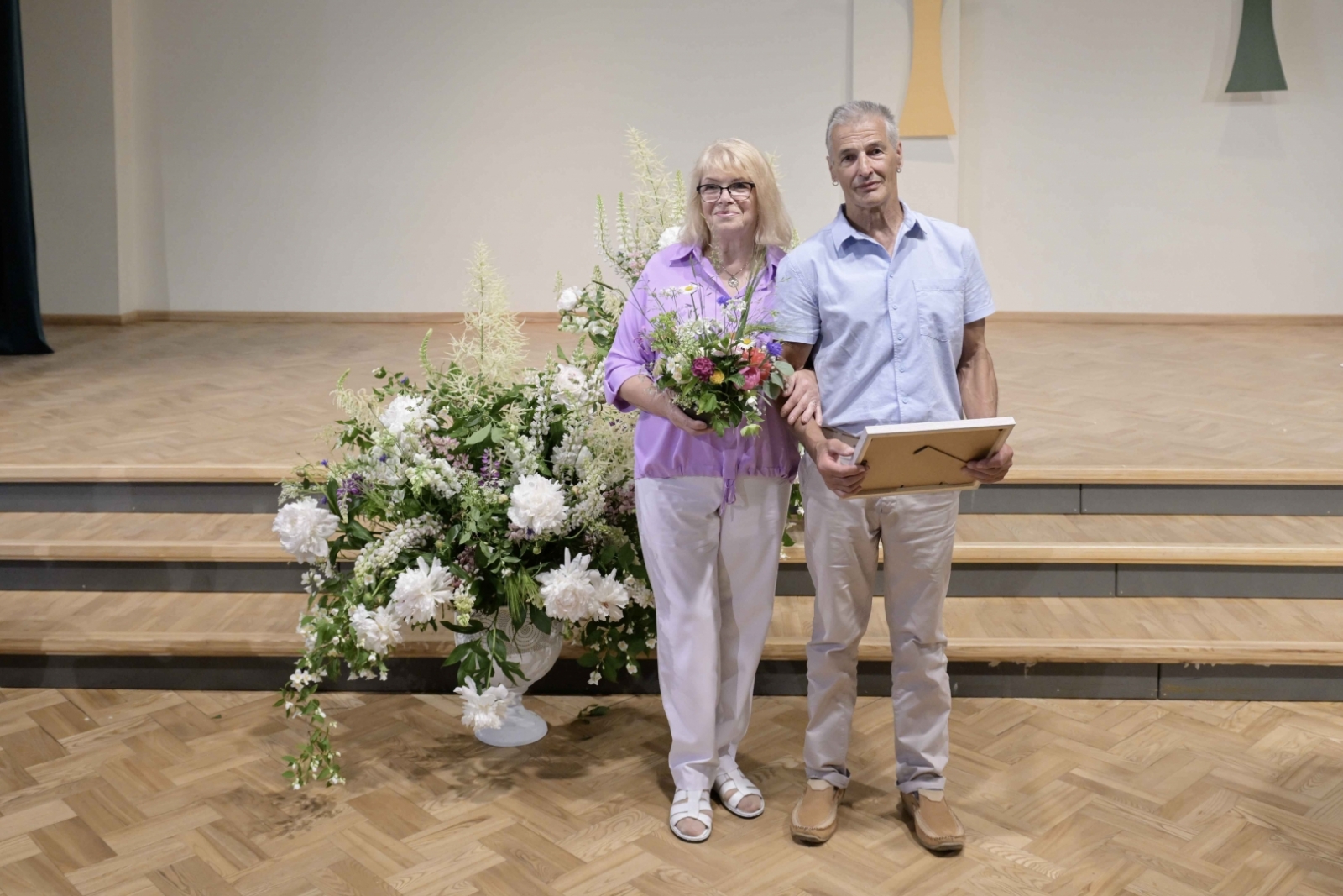 Vīrietis baltā kreklā, gaišās biksēs, sieviete rozā blūzē, gaišās biksēs Ogres novada Zelta kāzu pāris 31.05.2024