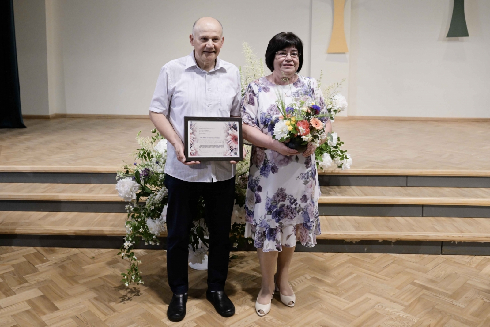 Sieviete kleitā ar ziediem, vīrietis gaišā kreklā, tumšās bikses ar pateicības rakstu Ogres novada Zelta kāzu pāris 31.05.2024