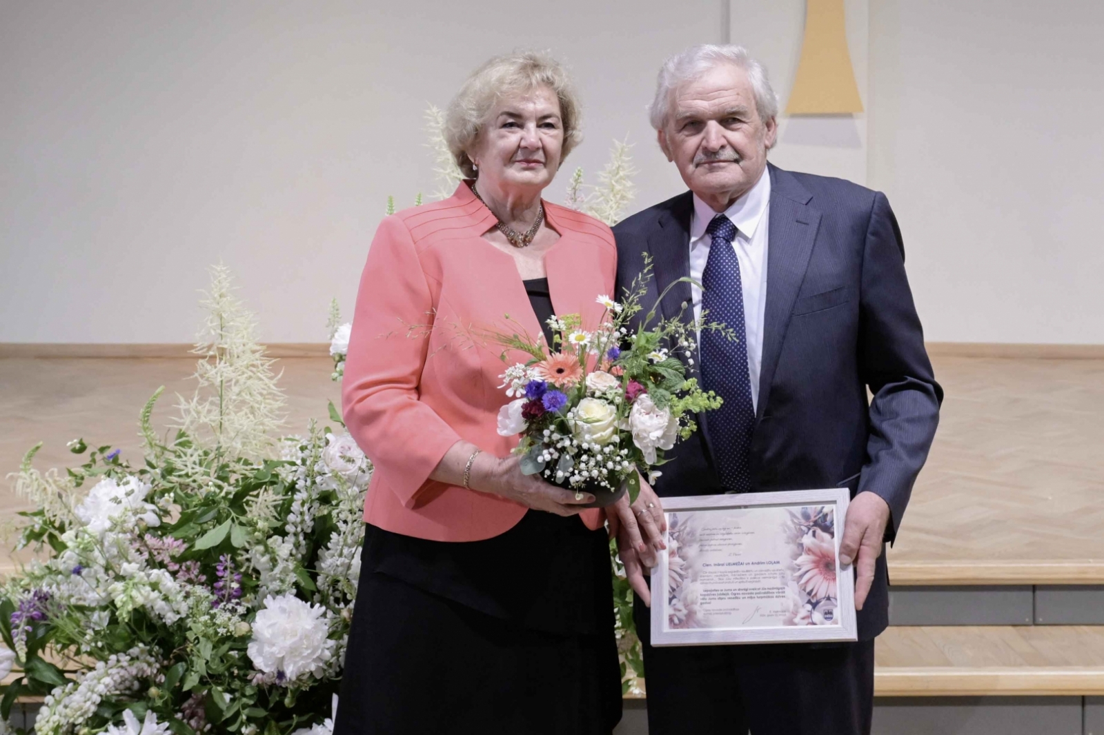 Sieviete melnā kleitā, rozā žaketē ar ziediem, vīrietis uzvalkā ar pateicības rakstu Ogres novada Zelta kāzu pāris 31.05.2024