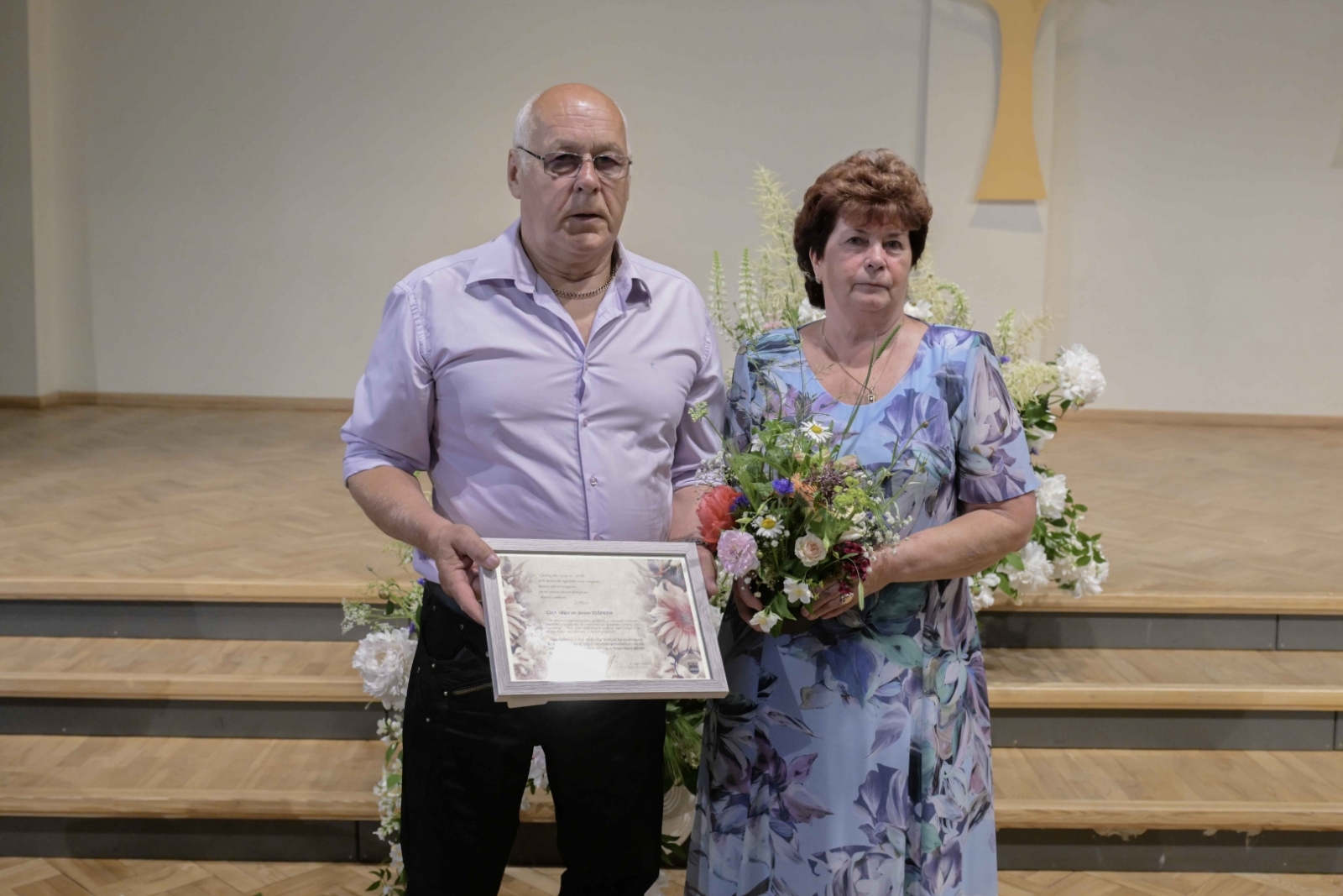 Vīrietis gaišā kreklā ar pateicības rastu, sieviete zilganā tērpā ar ziediem Ogres novada Zelta pāri 31.05.2024.