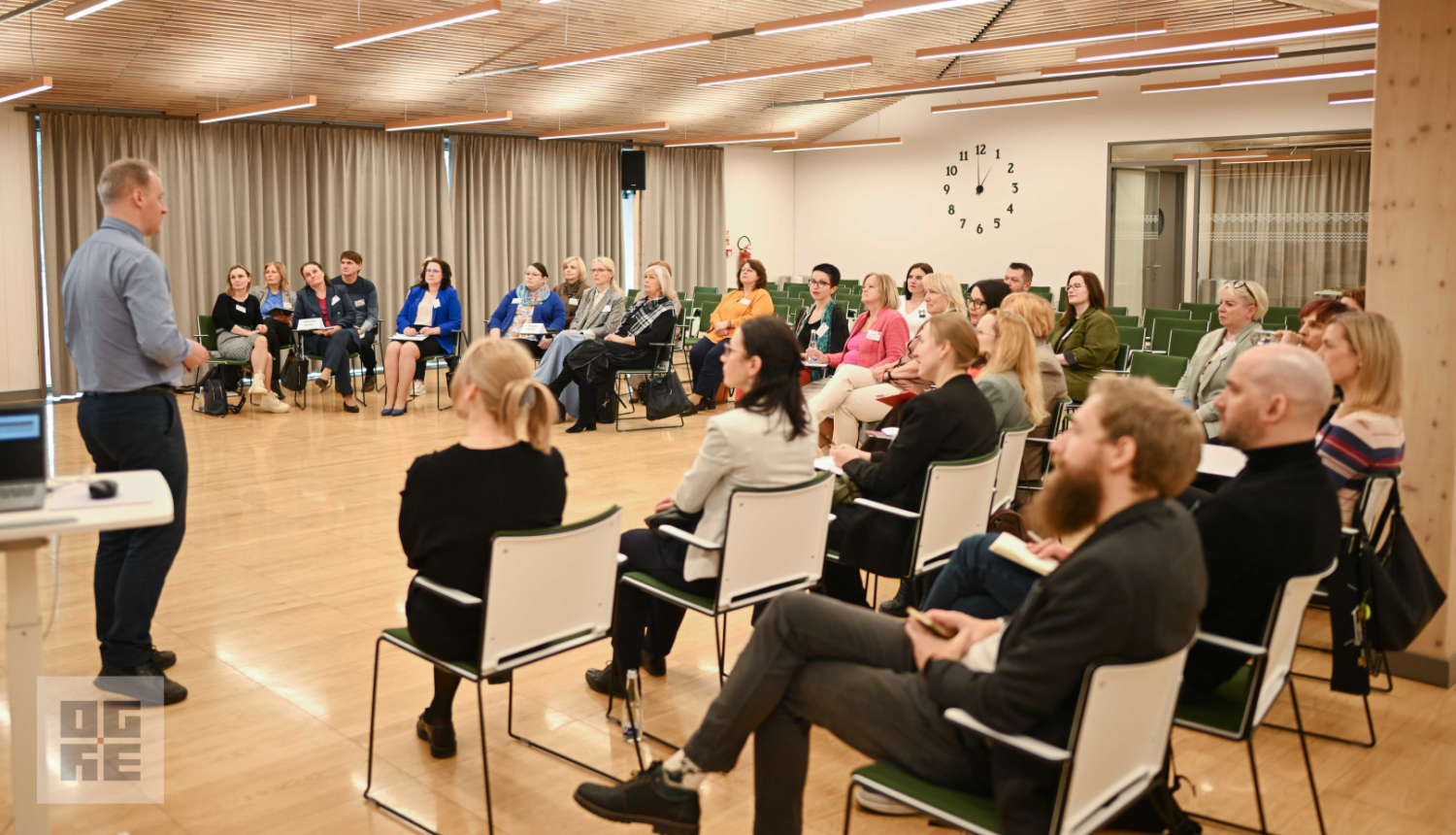 Tikšanās dalībnieki Ogres Centrālās bibliotēkas zālē, sēžot puslokā, klausās Ogres novada Izglītības pārvaldes vadītāja Igora Grigorjeva stāstījumu