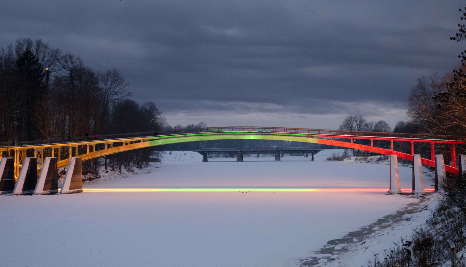 Lietuvas karoga krāsās - dzeltenā, zaļā un sarkanā - izgaismotais arkveida gājēju tilts pār aizsalušo Ogres upi