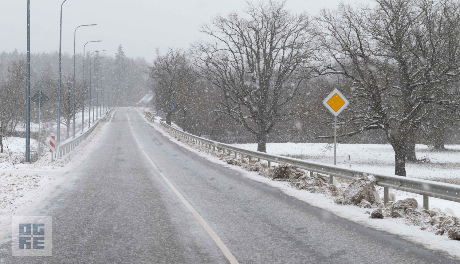 Asfaltēts ceļš Tīnūžos putenī ar norobežojošām metāla barjerām un ceļa zīmi "Galvenais ceļš"