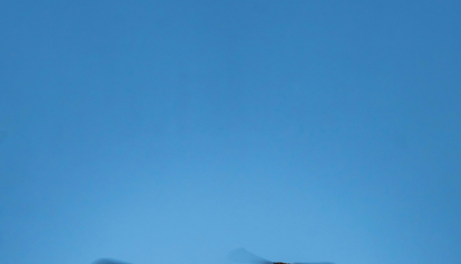 Attēls rakstam par iespējām apgūt dronu pilotēšanu. Attēlā lidojošs drons uz zilas debess fona