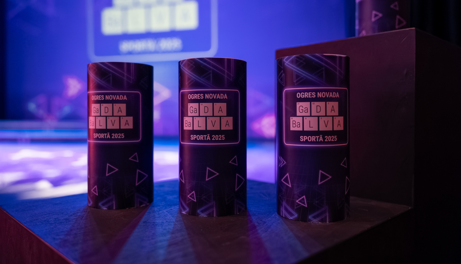 Uz podesta novietotas 3 balvas - tumši zili cilindri ar uzrakstu "OGRES NOVADA GaDA  BaLVA SPORTĀ 2025"