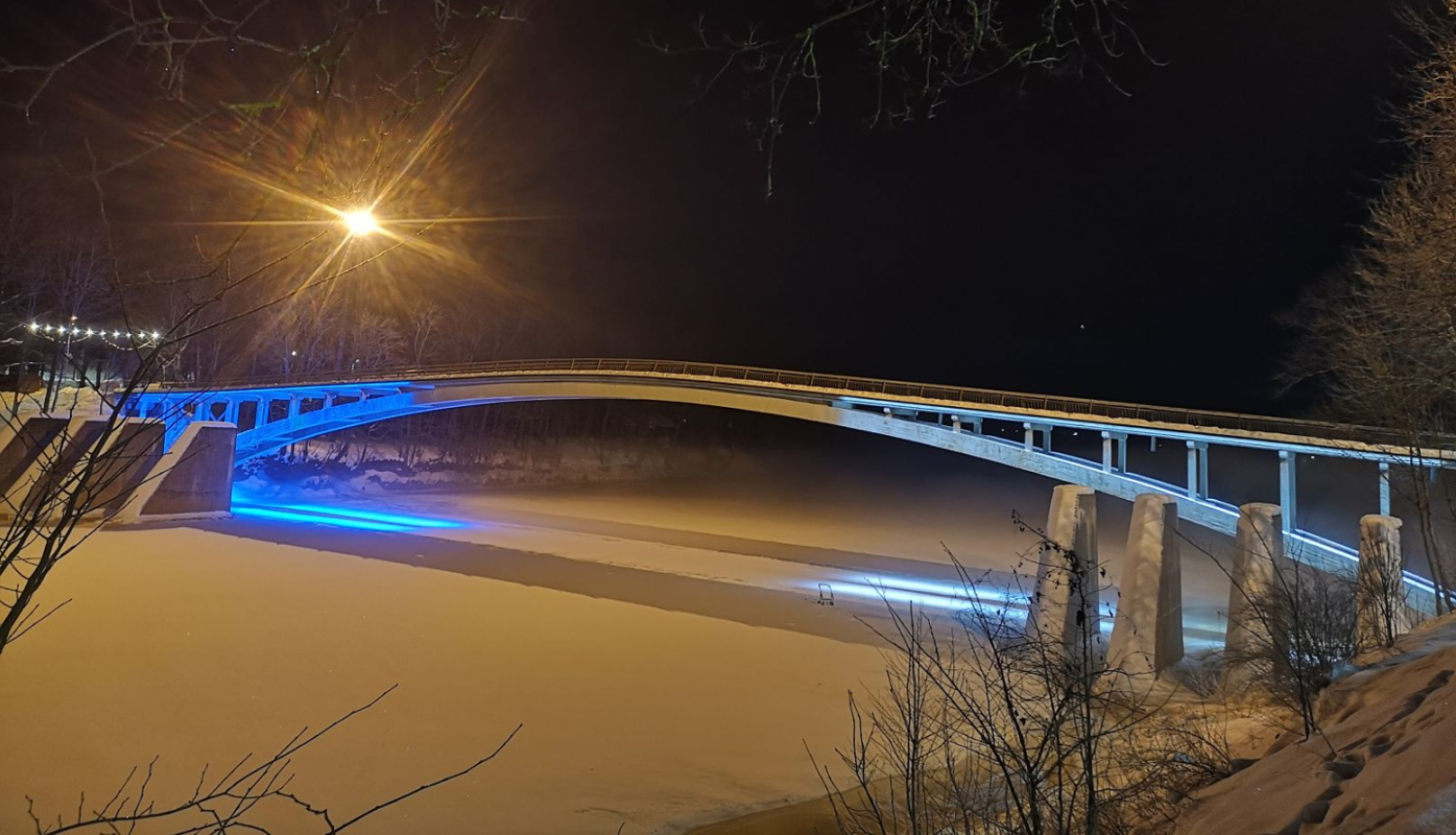 Igaunijas karoga krāsās izgaismotais arkveida gājēju tilts pār aizsalušo Ogres upi