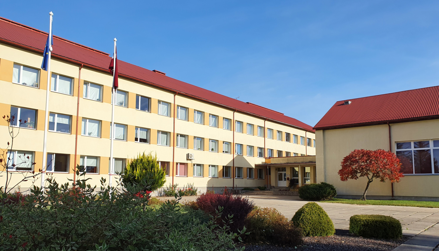 Skolas ēka ar diviem karogu mastiem un dekoratīvajiem stādījumiem priekšplānā