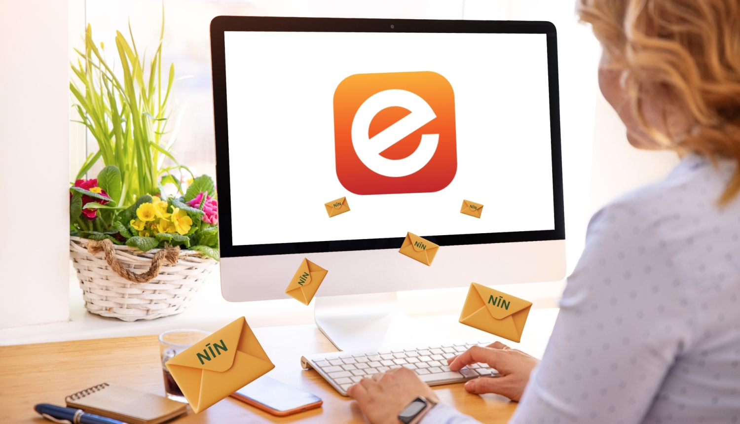 Attēlā ekrāns ar e-adreses simbolu - burtu e, pie ekrāna sēž sieviete, kas drukā uz klaviatūras. Apkārt lido aploksnes ar saīsinājumu NĪN (nekustamā īpašuma nodoklis). Uz palodzes aiz ekrāna groziņš ar pavasara ziediem.