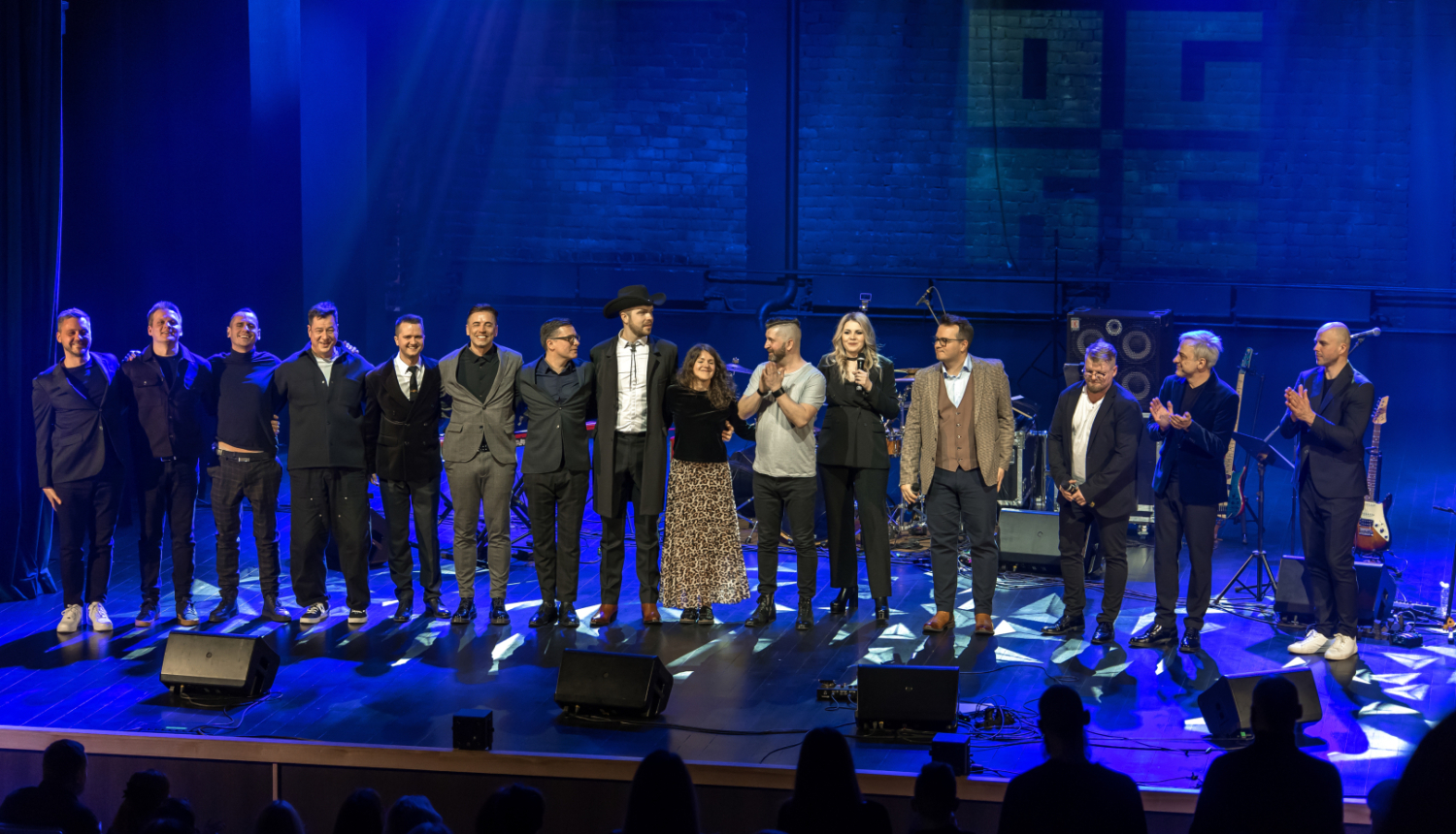 Skatuves mākslinieku kopbilde - 15 mūziķi koncerta noslēgumā nostājušies vienā taisnā līnijā uz Ogres novada Kultūras centra skatuves