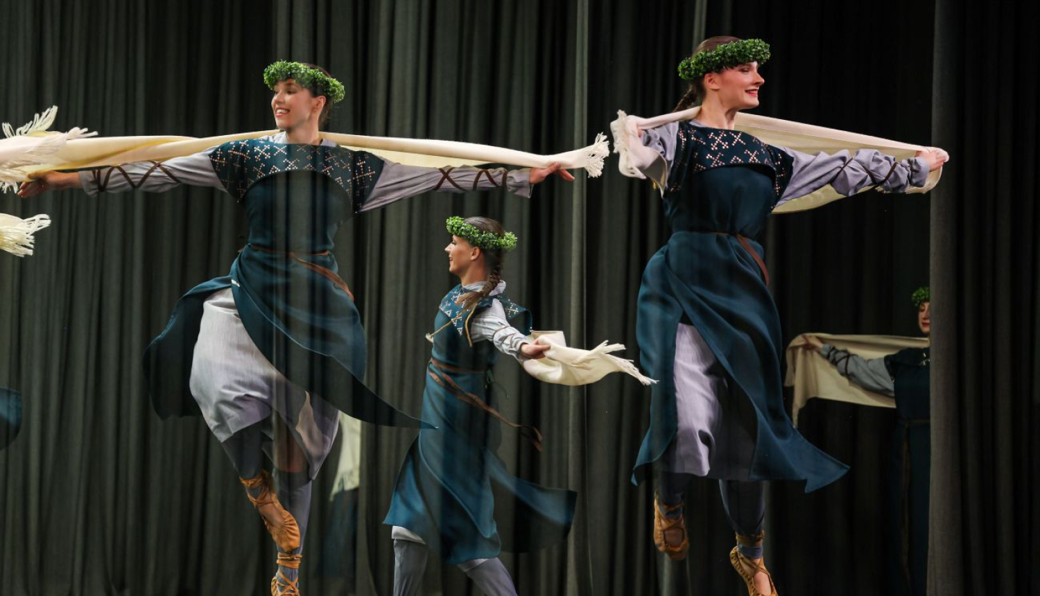 Dejojošas jaunietes zili baltos tērpos, pastalās un ar zaļiem vainagiem