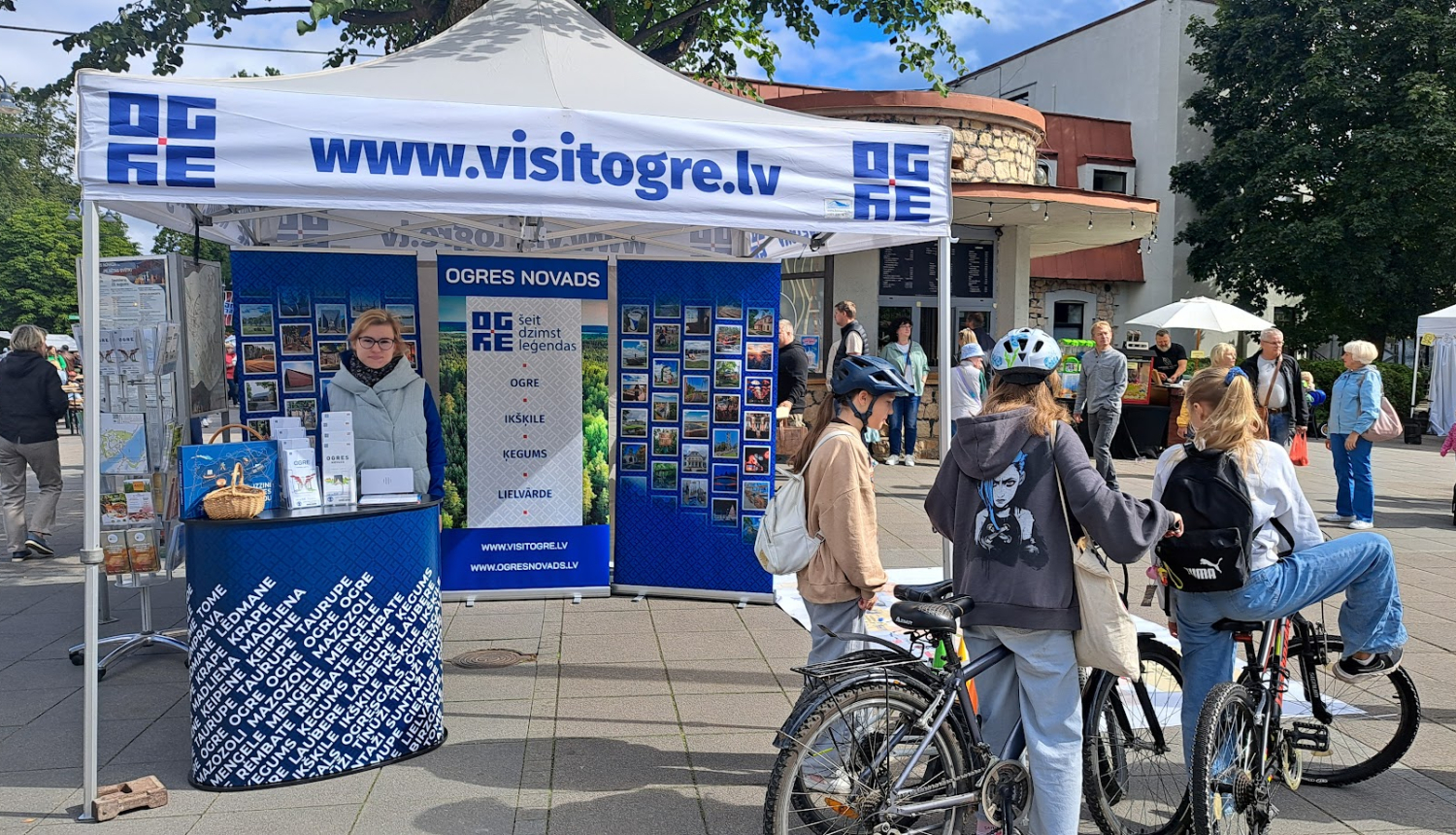 Visit Ogre tūrisma informācijas stends pasākuma laikā Ogres pilsētvidē. Stendā atrodas darbinieks. Priekšplānā jaunieši uz velosipēdiem.
