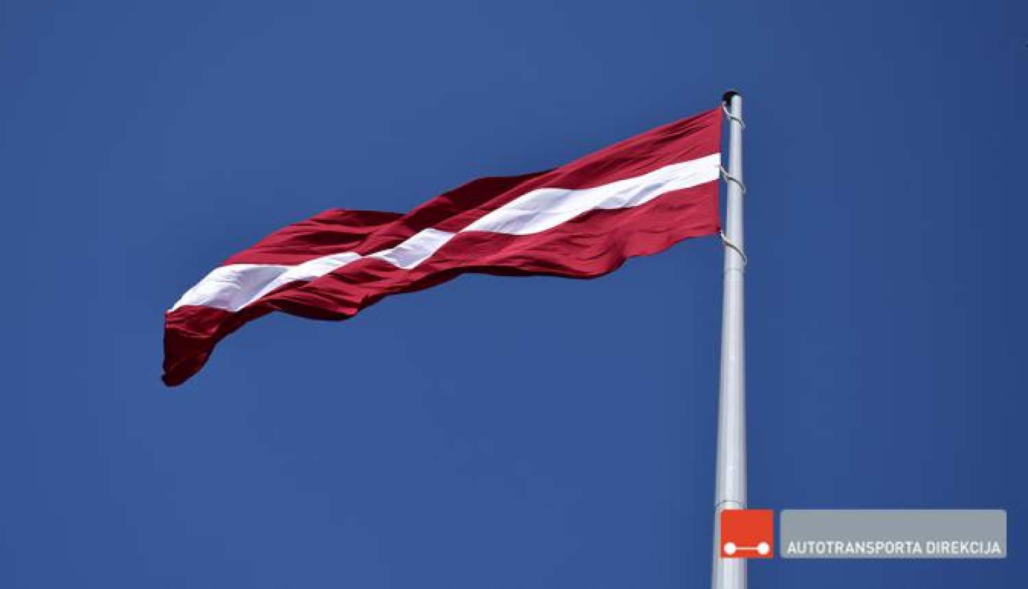 Latvijas Valsts sarkanbaltsarkanais karogs plīvo mastā uz zilu debesu fona, Autotransporta direkcijas logo uz attēla labajā apakšējā stūrī