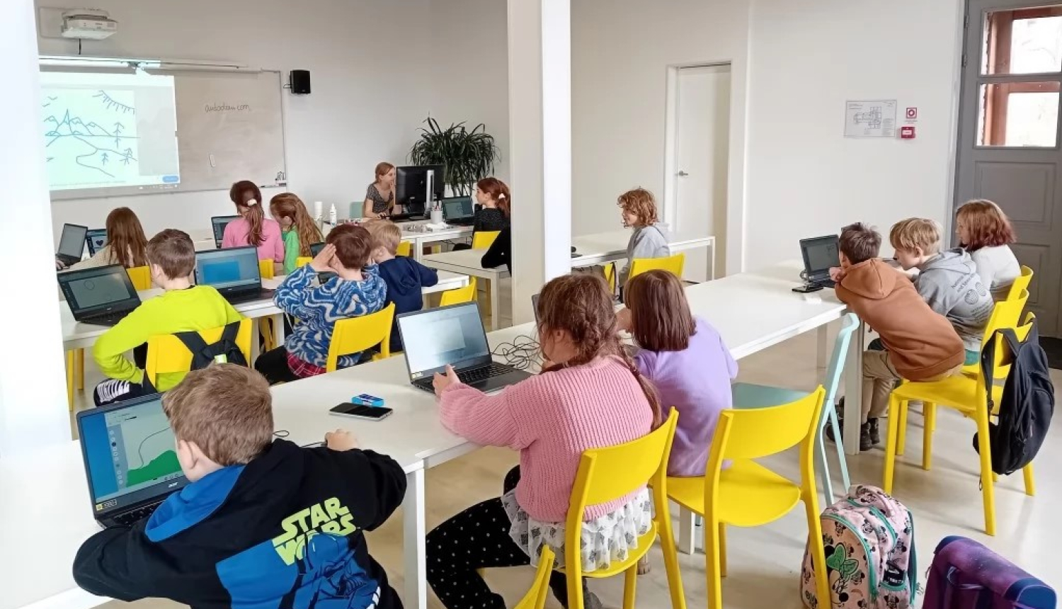 Aicinājums pieteikties Rīgas Tehniskās universitātes Bērnu un jauniešu universitātes interešu izglītības programmām Ogrē. Attēlā bērni sēž pie galdiem uz dzelteniem krēsliem. Viņiem priekšā digitālā tāfele