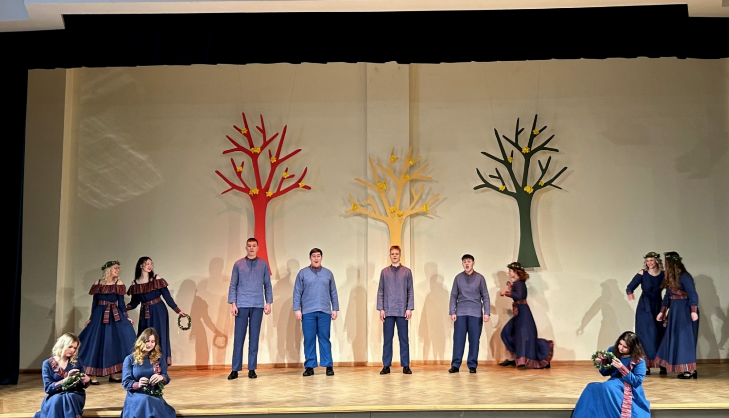 Jaunieši zilos tērpos dzied uz skatuves, fonā dekorācijās krāsaini koki 02.2024.