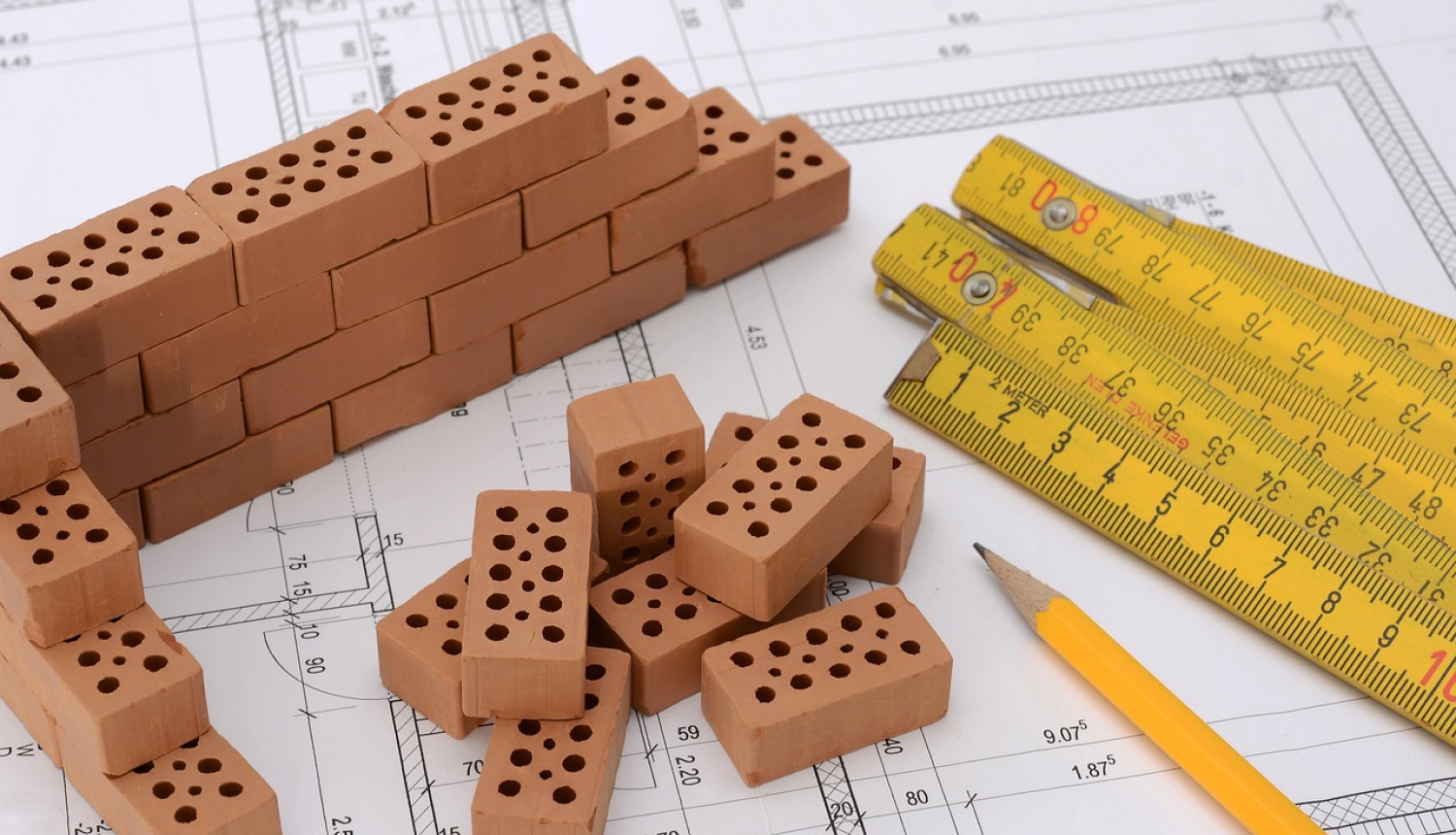 Būvniecības darbi Ogres novadā. Attēlā rasējums mērlenta, zīmulis un ķieģeļi