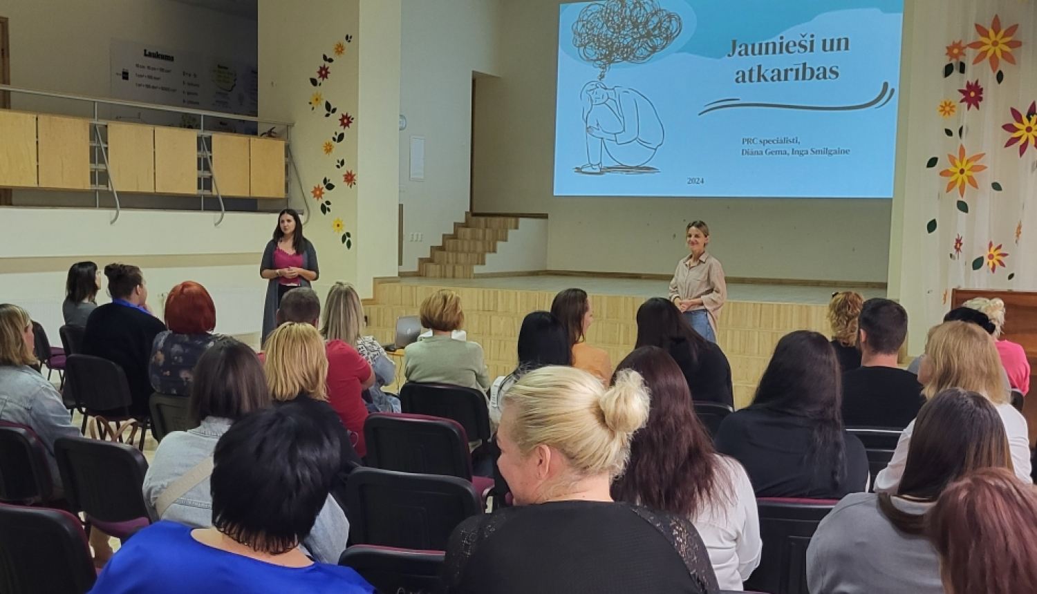 Cilvēki redzami ar mugurām, sēž krēslos, priekšā pasniedzējs stāsta par atkarību izraisošām vielām. 25.09.2024.