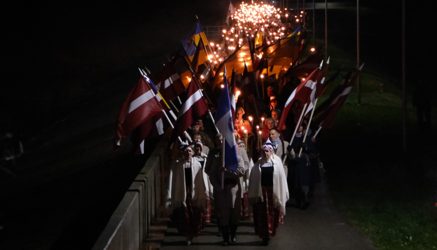 Cilvēki, priekšplānā tautas tērpos, dodas gājienā ar Latvijas karogiem un lāpām pa Ogres molu par godu Lāčplēša dienai