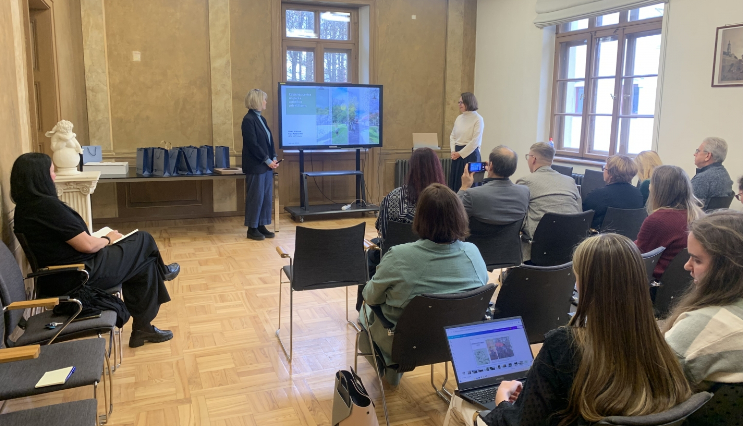 Pasākuma norises zāles kopskats ar dalībnieku sēdvietu rindām, ekrānu un divām studentēm, kas sniedz prezentāciju