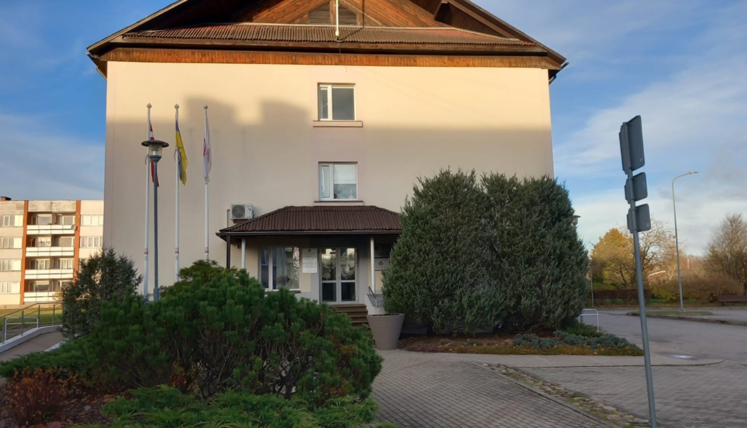 Lielvārdes pilsētas un pagasta pārvaldes ēka ar karogu mastiem un skujeņu stādījumiem priekšplānā