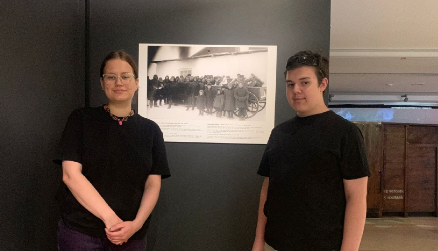 2 cilvēki melnos kreklos, gaišās biksēs stāv pie melnbaltas fotogrāfijas Ogres Vēstures un mākslas muzejā