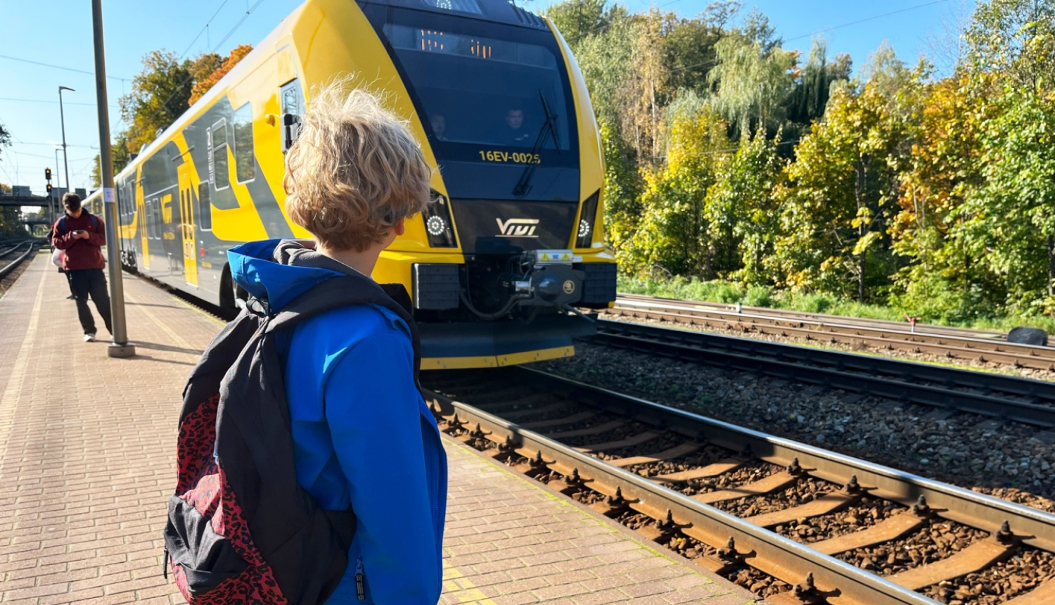 Jaunietis zilā jakā ar mugursomu stāv pie vilciena