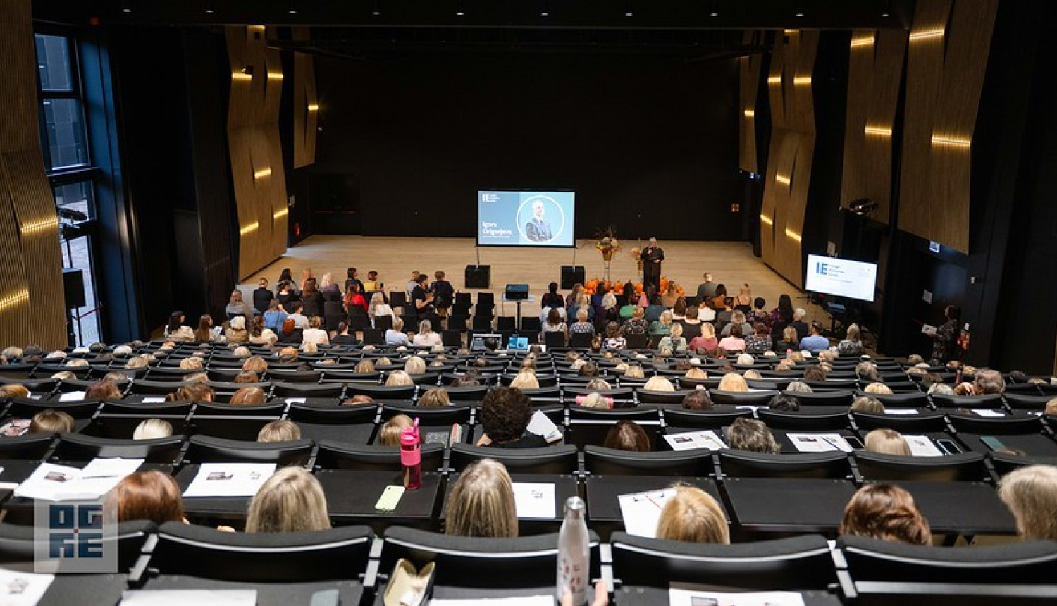 Ogres Valsts ģimnāzijas auditorijā sapulcējušies novada pedagogi uz konferenci. Fonā redzama prezentācija.