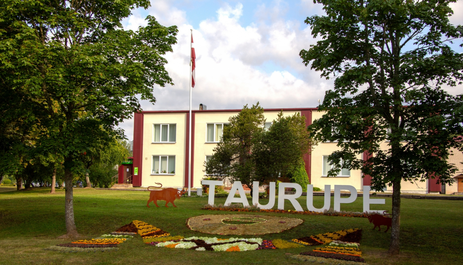 Uzraksts Taurupe ar ziediem, fona daudzstāvu māja Taurupes pagasta svētkos 07.2024.